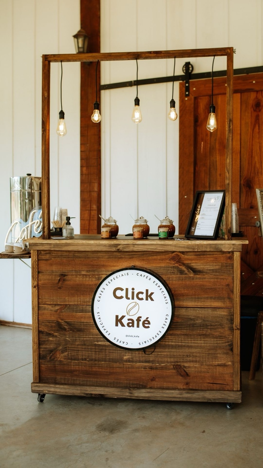 Estação Raízes - Click Kafé com madeira escura, lâmpadas Edison e logo redondo na frente